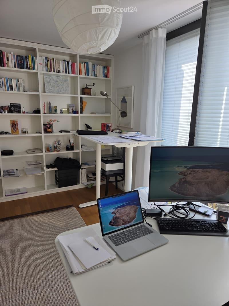 Furnished Room. A well-organized home office with a white desk, laptop, large screen, keyboard, and a bookshelf filled with books and decorative items.