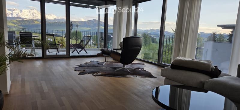 Furnished Room. Modern living room with large windows offering a scenic mountain view, featuring a sofa, chairs, coffee table, and a small table on the balcony.