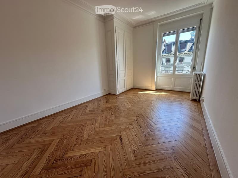 Empty room with wooden herringbone flooring, white walls, and large window with view of rooftops.