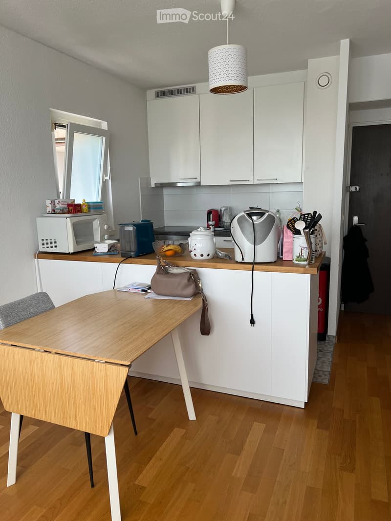 Modern kitchen with white cabinets, wooden countertop, microwave, electric kettle, and a small wooden table with a chair.