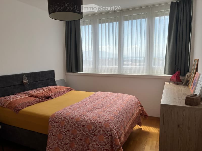 A cozy bedroom with a large window, a bed with a colorful bedspread, a nightstand with a lamp, and decorative items including a picture frame and a small toy.