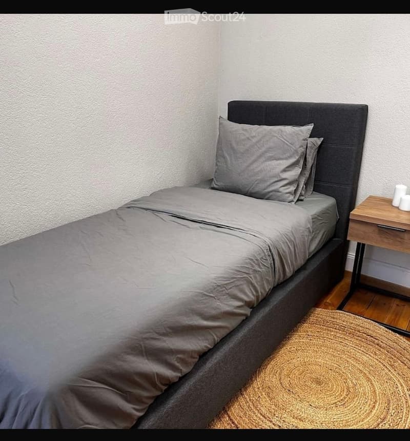 A small, neatly arranged bedroom with a single bed, grey bedding, a wooden nightstand, and a woven area rug on the floor.
