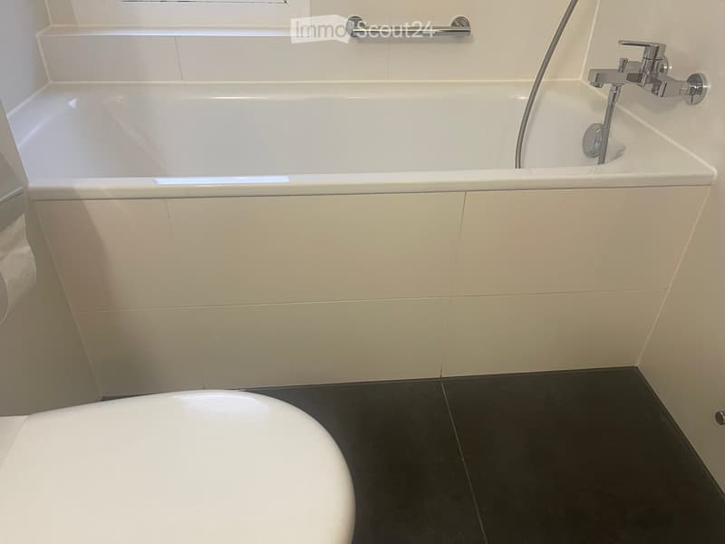 Modern bathroom with a white bathtub, chrome faucet, and handrail above the tub. The walls are tiled in light beige, and the floor is dark grey.