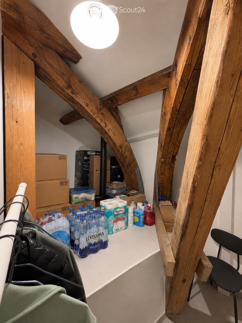 Attic storage area with exposed wooden beams, a hanging light fixture, a chair, water bottles, cleaning supplies, and cardboard boxes.