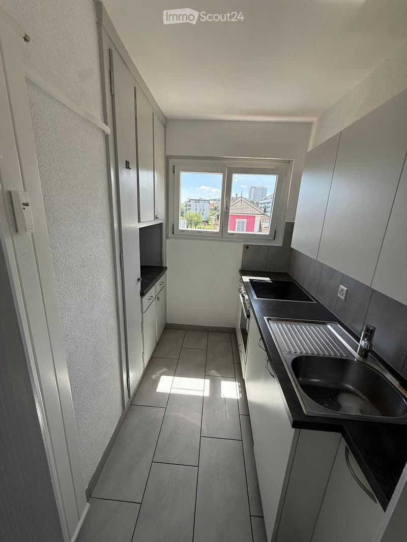 Modern kitchen with white cabinets, dark countertops, and a stainless steel sink. The space features tiled flooring and a window offering a view of the city.