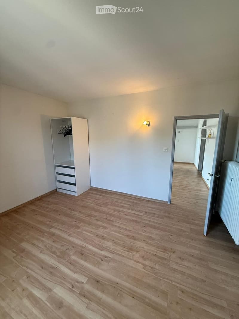 Empty room with light wooden flooring, white walls, and a built-in closet with drawers and hanging space. A wall-mounted light fixture is visible, and an open doorway leads to another area.