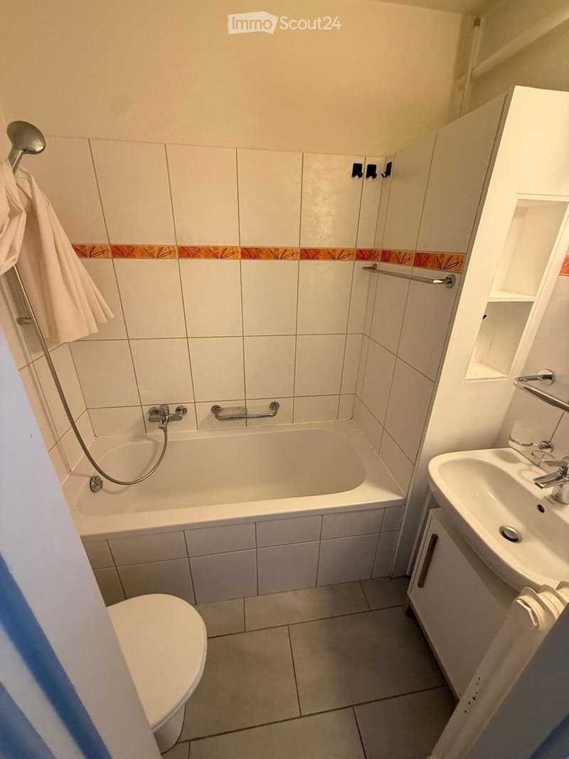 A compact bathroom with a bathtub, toilet, sink, and towel hanging on the wall. The walls are tiled with white tiles and a decorative border.