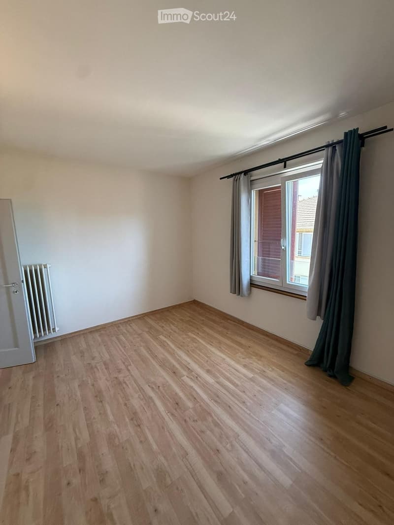 Empty room with light wooden flooring, white walls, a window with dark curtains, and a radiator on the left side.