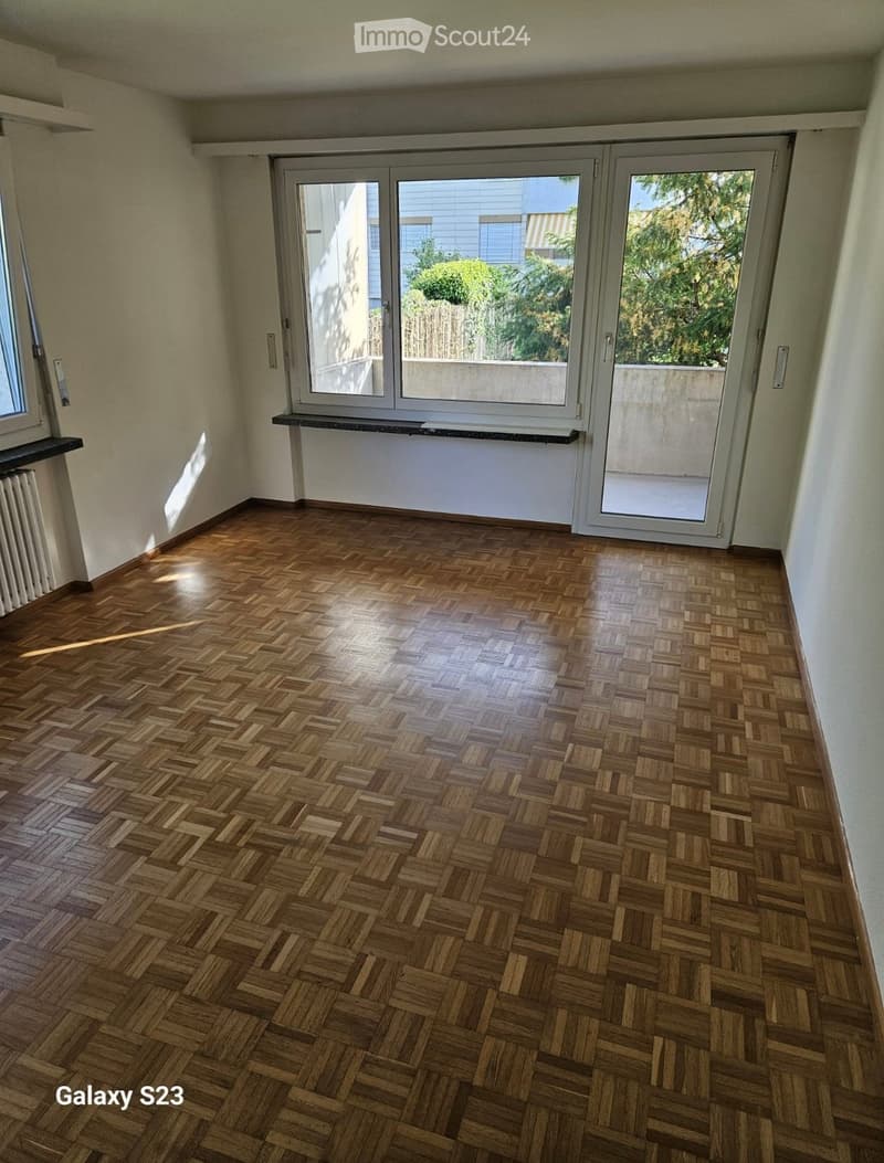 Empty room with wooden herringbone flooring, large windows, and sliding glass doors leading to a balcony.