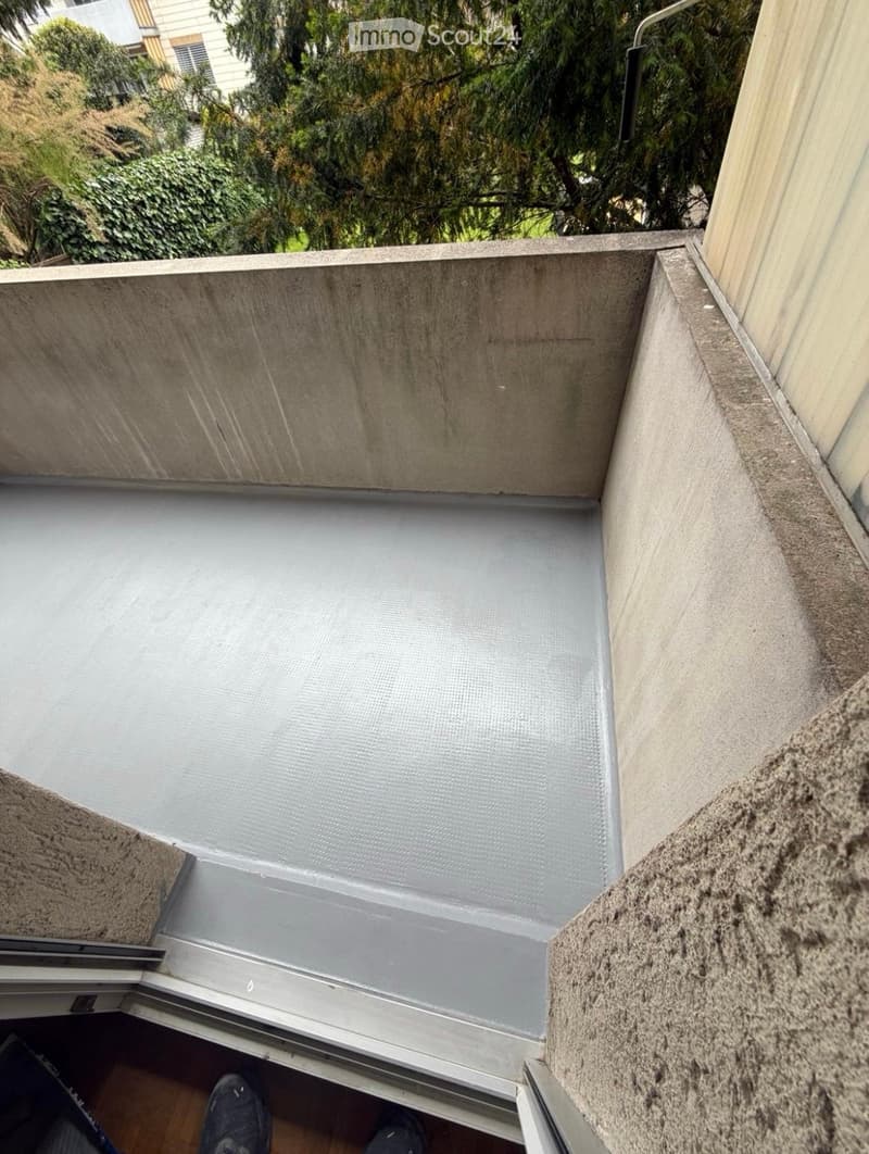 A small, enclosed balcony with a concrete floor and walls, featuring a metal safety railing and a sliding glass door leading to the interior.