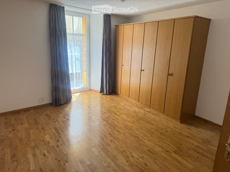 Empty room with wooden flooring, large window with curtains, and built-in wooden wardrobe.
