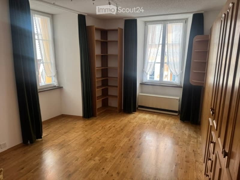 Empty room with wooden flooring, large windows with dark curtains, a radiator, and a wooden bookshelf.
