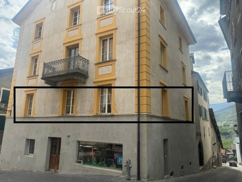 Multi-story building with yellow-framed windows and balconies, located in a European town with a visible street and storefront below.