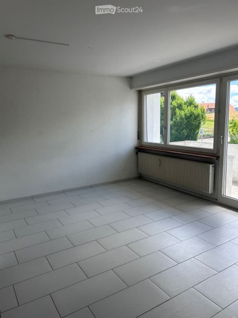 Empty room with white tiled floor, white walls, large window with wooden frame, radiator below window, and ceiling light fixture.
