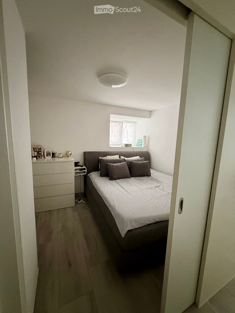 Cozy bedroom with a double bed, nightstand, lamp, and picture frame on the dresser. The room has light-colored walls and a window with a view.