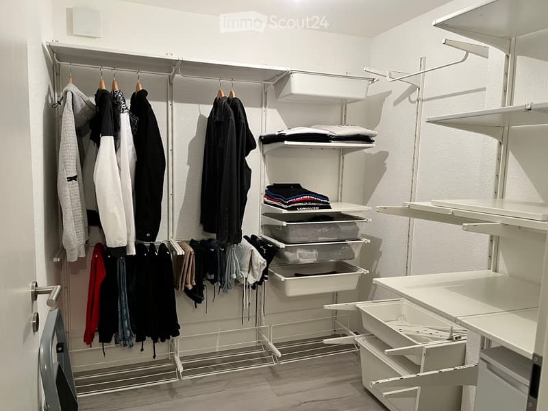 A modern, well-organized closet with white walls, wooden flooring, and multiple storage units including shelves, drawers, and hanging space for various clothing items.