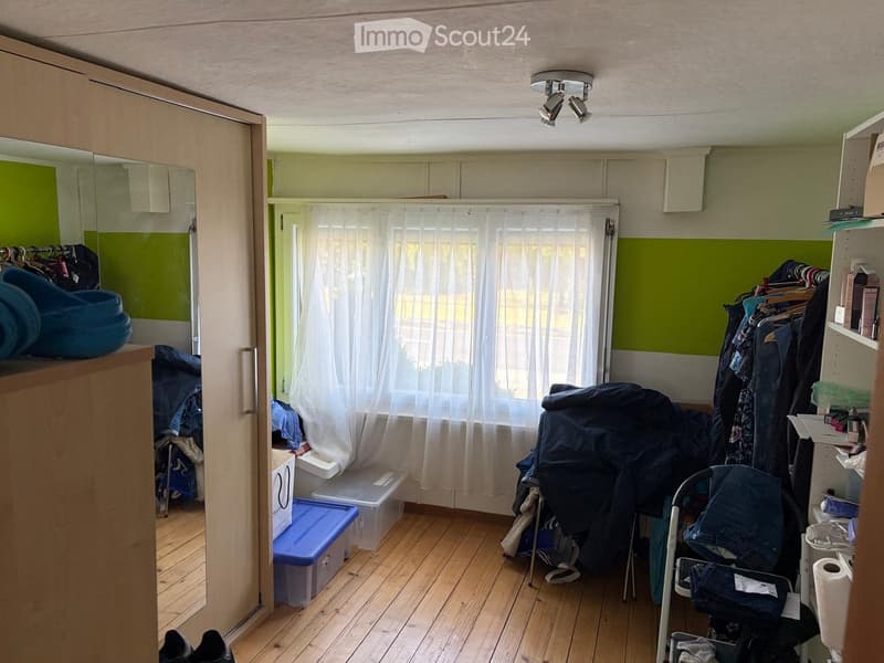 A small, furnished room with wooden flooring, green walls, a large window with white curtains, a wardrobe with hanging clothes, a mirror, storage boxes, and a baby stroller near the window.
