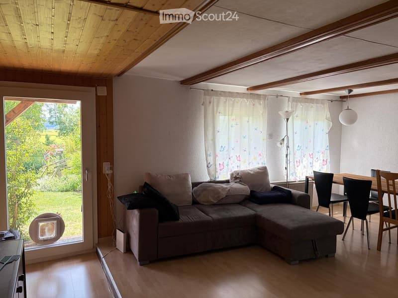 Bright living room with wooden floors, exposed wooden beams, large windows with sheer curtains, a grey sectional sofa, a dining table with chairs, and a floor lamp.