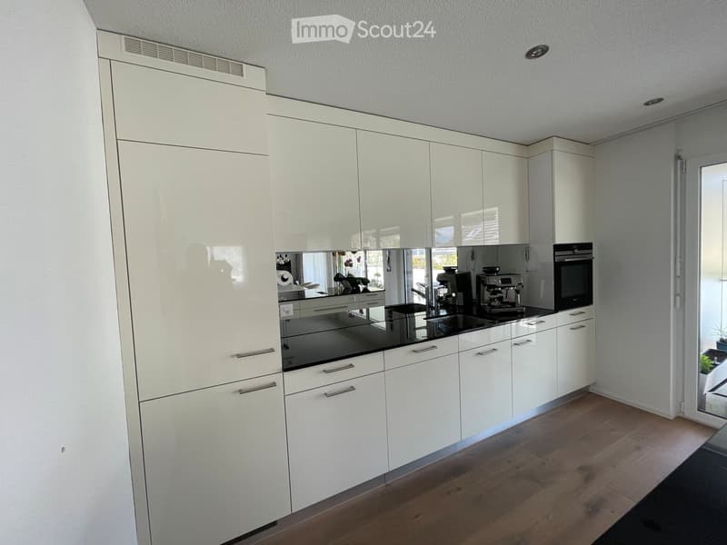 Modern kitchen with white cabinets, black countertops, and stainless steel appliances including a coffee maker and oven. Wooden flooring and large windows provide natural light.