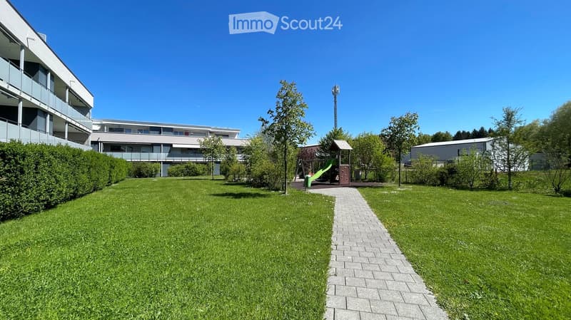 Well-maintained garden area with green grass, a paved walkway, a small play structure, and surrounding buildings with balconies.