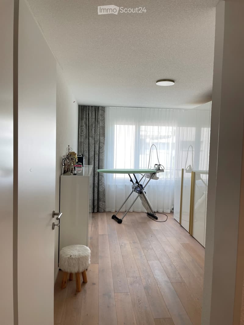 A small, well-lit room with wooden flooring, a white fur-covered stool, an ironing board, a mirror, and a curtained window.
