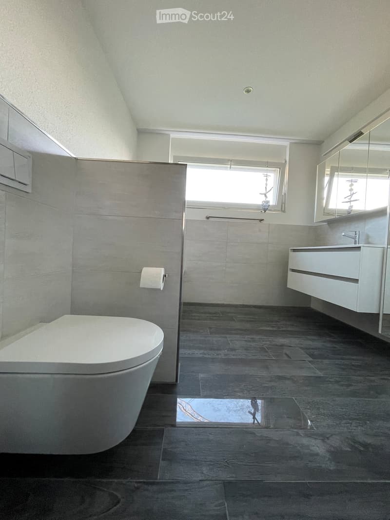 Modern bathroom with a toilet, sink, mirror, and window. Features dark tiled flooring and light grey walls.