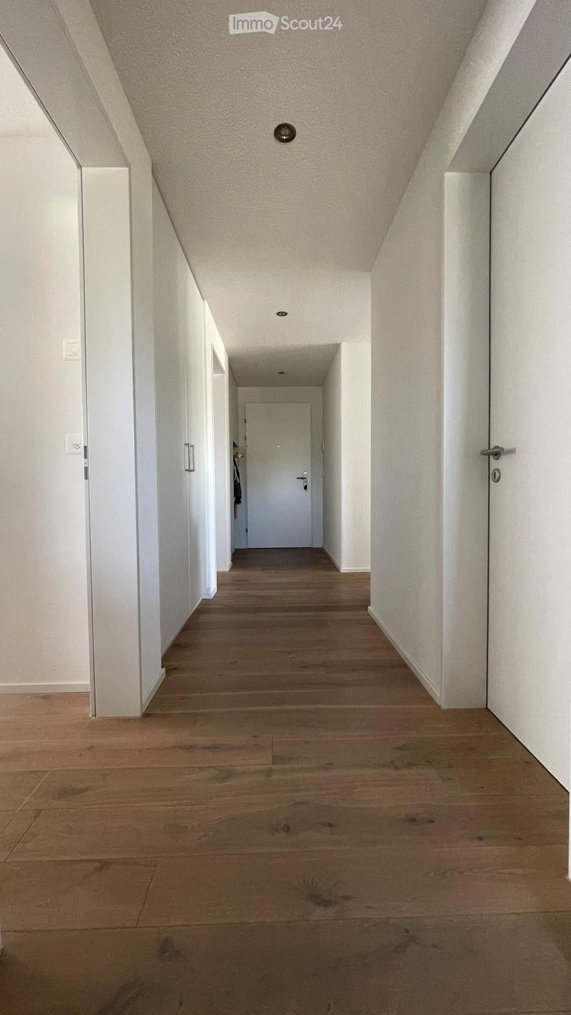 A long, narrow hallway with wooden flooring and white walls, featuring several doors and recessed lighting.