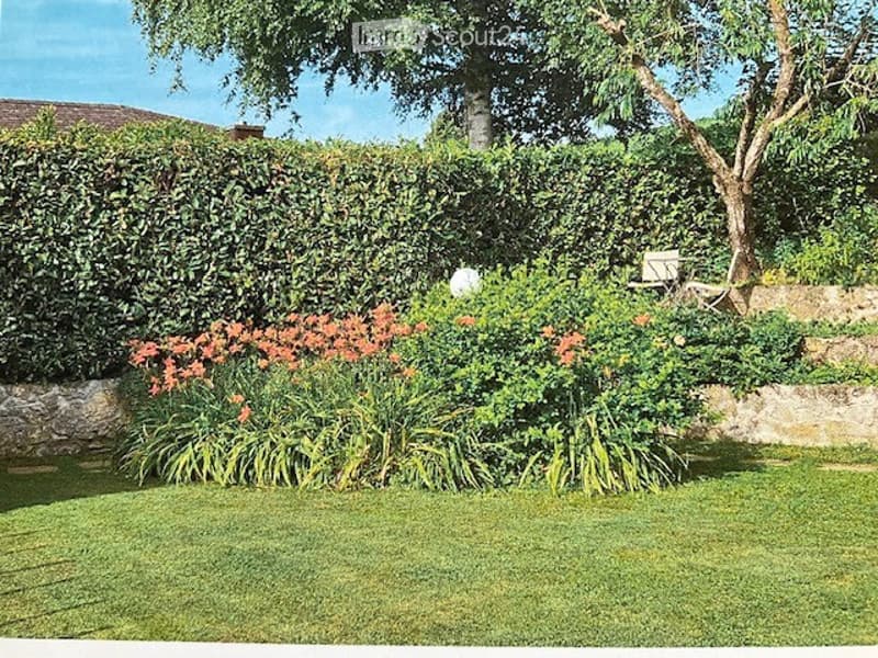 Well-maintained garden with a neatly trimmed hedge, vibrant orange flowers, and a tree providing shade. The lawn is lush and green, bordered by a stone wall.