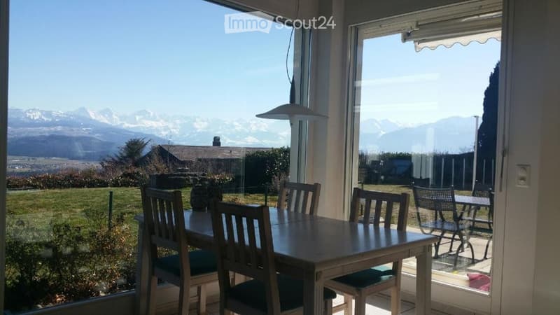 Bright dining area with a wooden table and chairs, large windows offering a scenic mountain view, and a small outdoor seating area visible through the glass door.