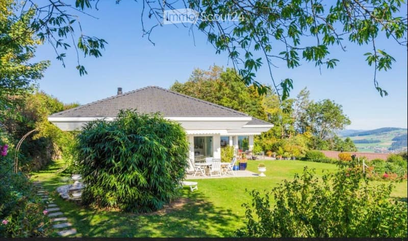 A white house with a tiled roof is surrounded by a well-maintained garden, featuring a patio area with outdoor furniture and a scenic view of the countryside.