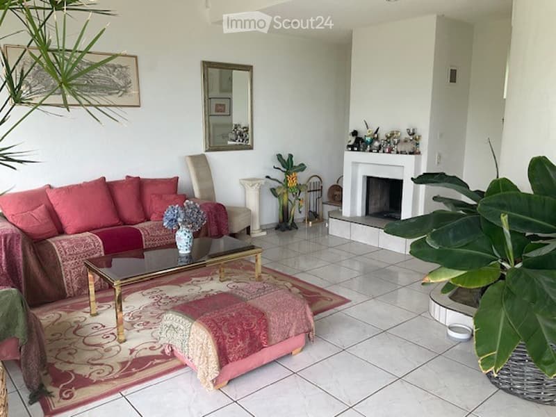 Bright living room with white tiled flooring, red sofa, coffee table, ottoman, fireplace, and decorative plants.