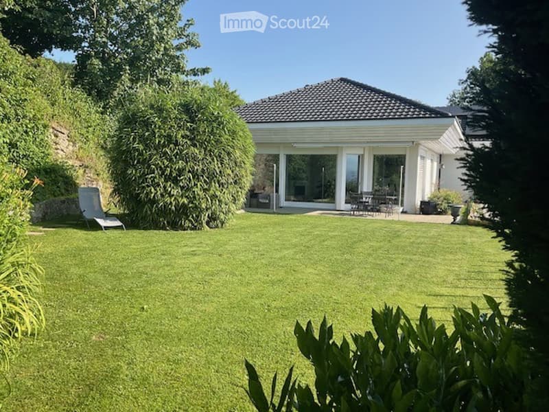 A well-maintained garden with a modern white house in the background, featuring a patio with outdoor furniture, a lounge chair, and lush greenery.