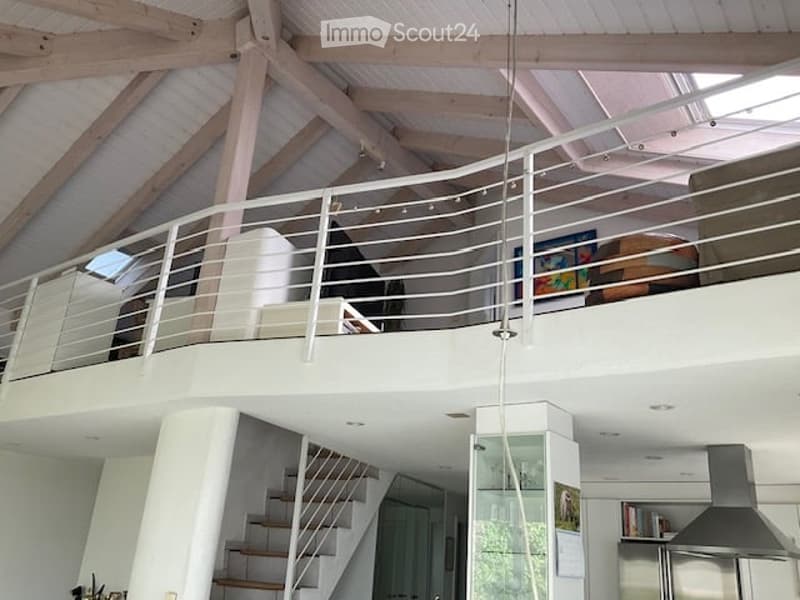 Open-plan living space with a raised ground floor, exposed wooden beams, and a modern balcony overlooking the lower level. Features a sofa, table, chair, bookshelf, and framed picture.