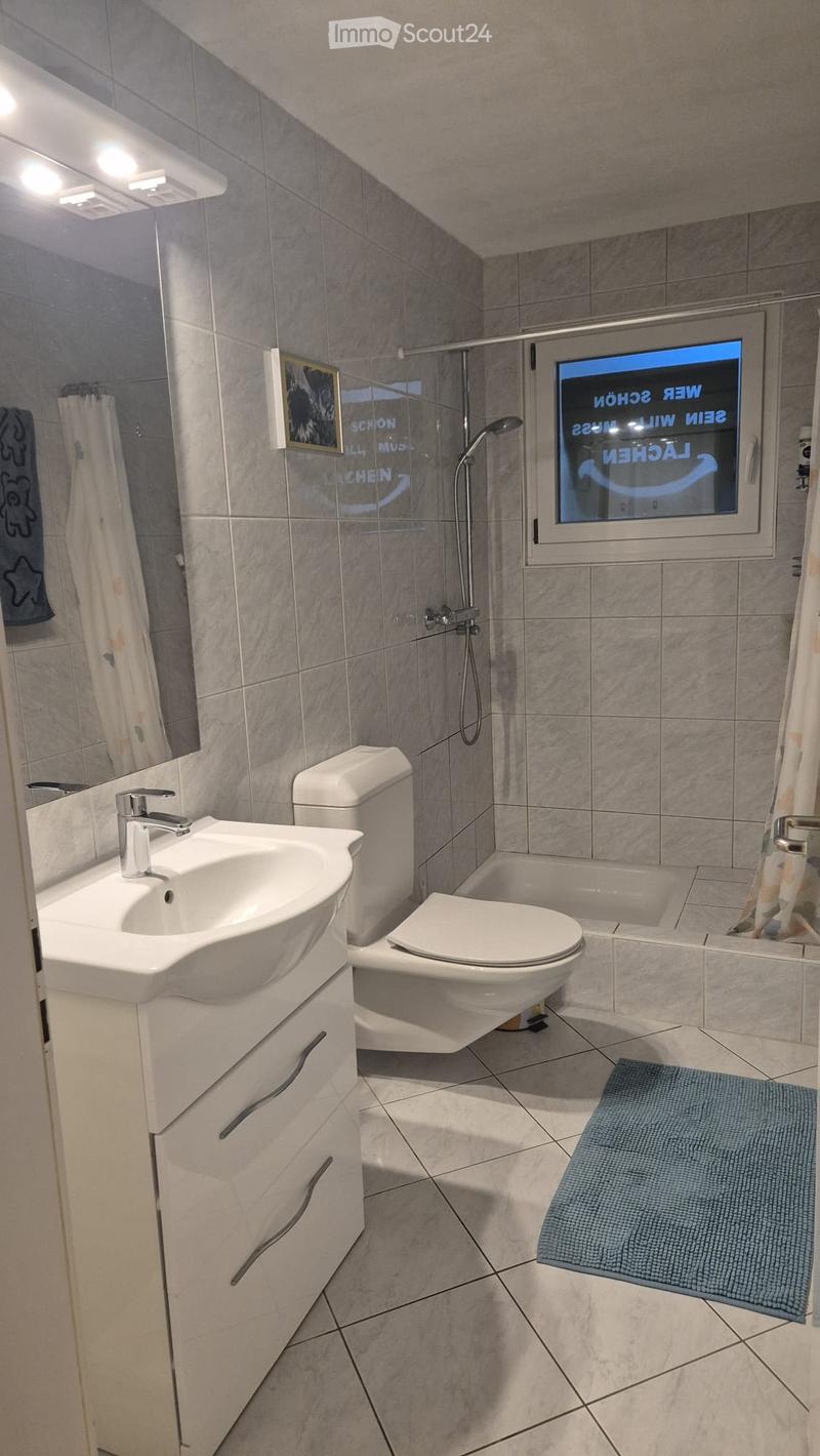 Modern bathroom with a white sink, toilet, bathtub, and shower area. Features a large mirror, modern faucet, and a blue bath mat on the floor.