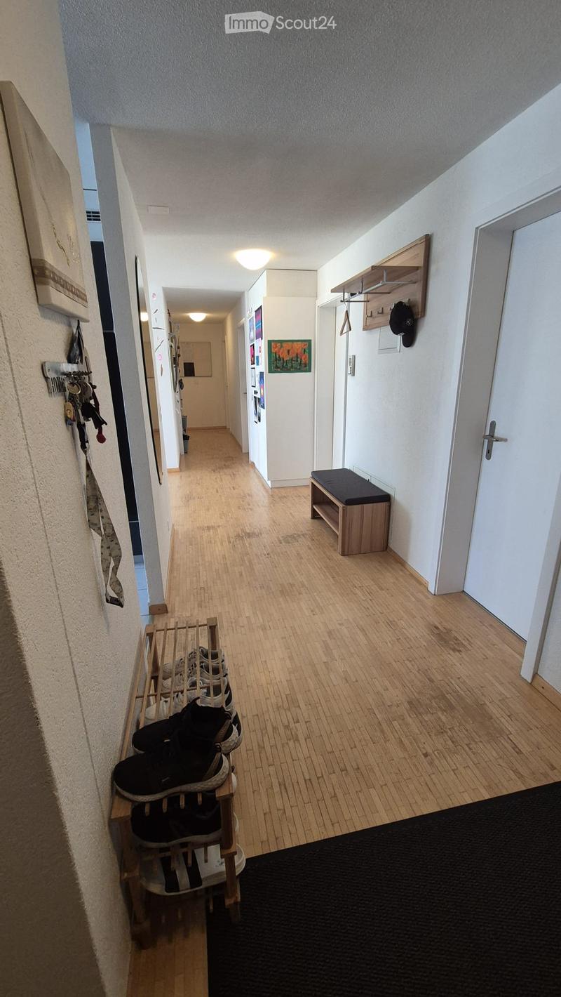 A well-lit hallway with wooden flooring, white walls, and several doors leading to different rooms. There are shoe racks with shoes, a small bench, and wall-mounted coat hooks with coats and hats.