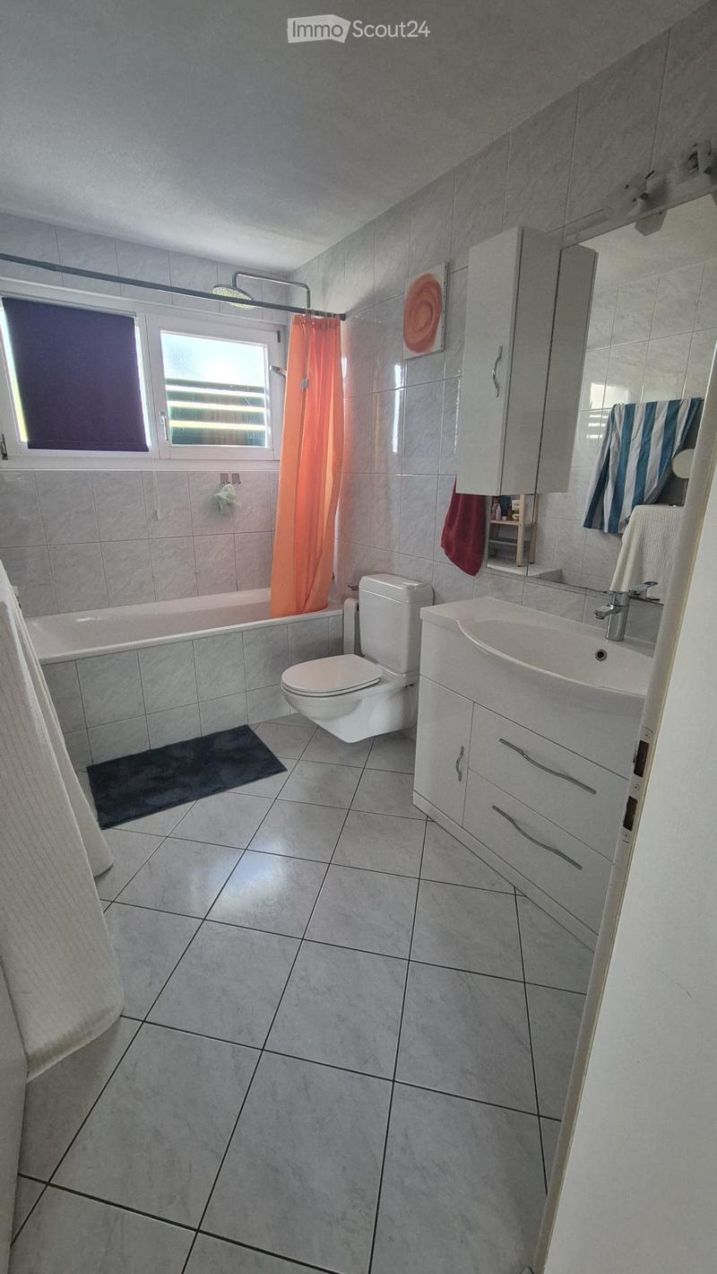 Modern bathroom with grey tiles, a bathtub, toilet, sink, mirror, and a small window with blinds.