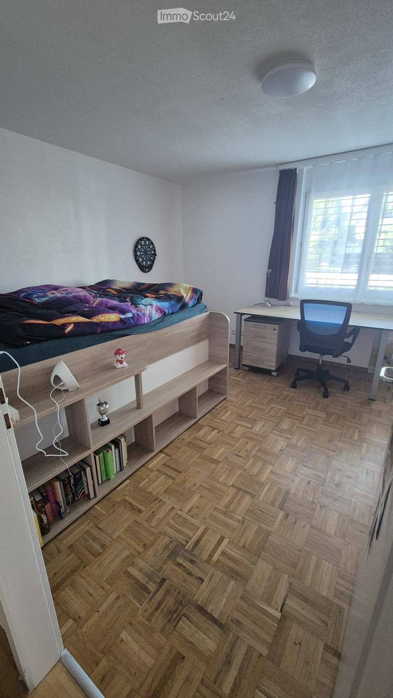 A well-lit bedroom with a lofted bed, a desk with a chair, a wooden shelf with books and a lamp, and a dartboard on the wall.