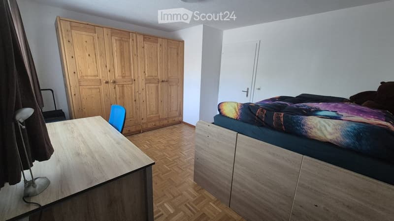 Bedroom with a bed, colorful bedspread, large wooden wardrobe, desk, chair, and lamp.