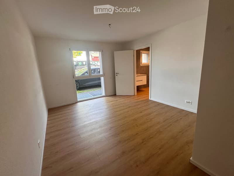 Empty room with wooden flooring, white walls, and a sliding glass door leading outside. A small adjacent room with a cabinet is visible through an open door.