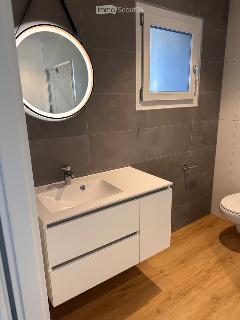 Modern bathroom with a white sink, chrome faucet, round illuminated mirror, and a toilet. Grey tiled walls and wooden flooring.