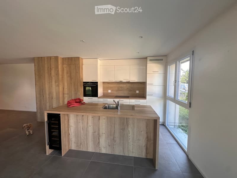 Modern kitchen with a wooden island, white cabinets, stainless steel sink, and built-in oven. A red bag rests on the island, and a small dog is visible in the background.