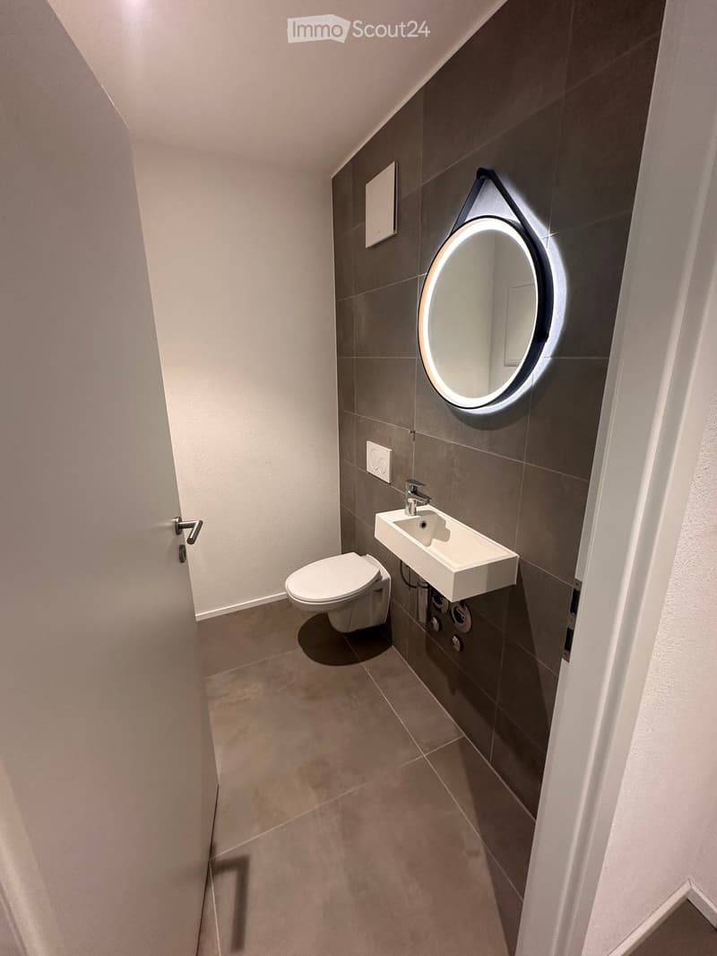 Modern bathroom with a toilet, sink, and illuminated round mirror. Grey tiled walls and beige tiled floor.