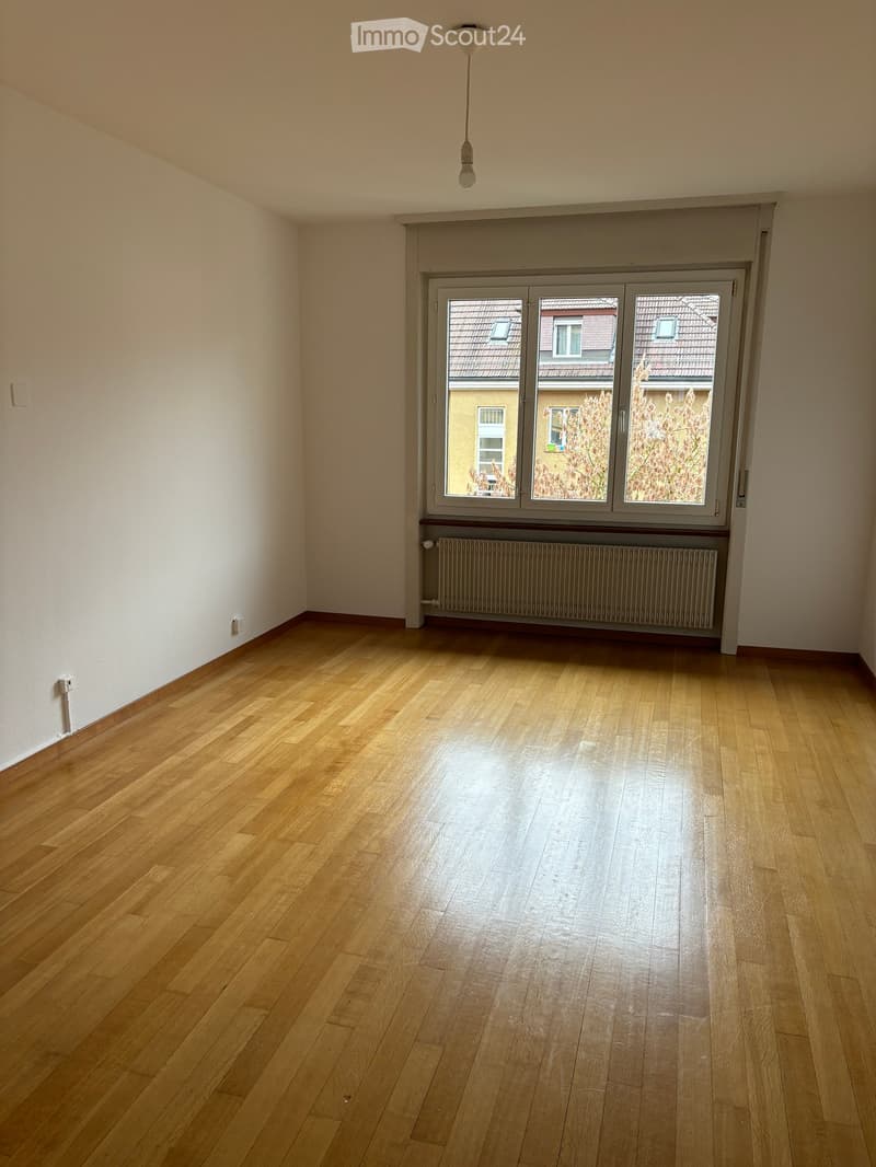 Empty room with wooden flooring, white walls, large window with view of neighboring buildings, radiator, and a hanging light fixture.