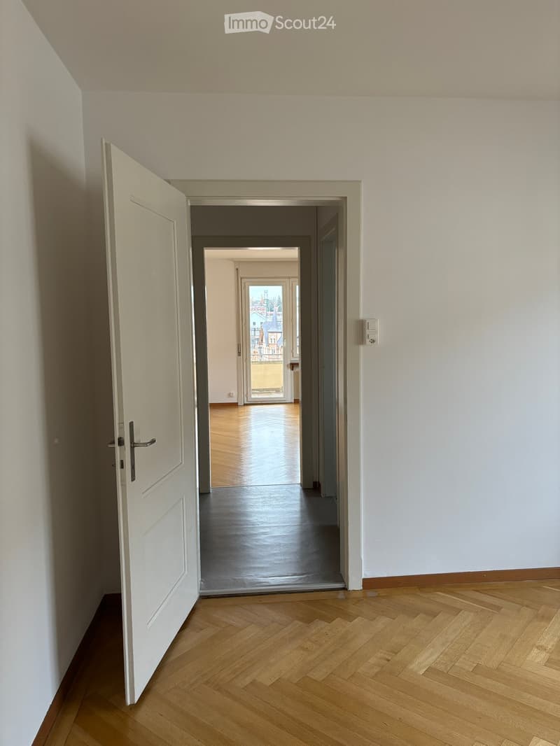 Empty hallway with wooden flooring, white walls, and an open door leading to another room with a view of a cityscape.