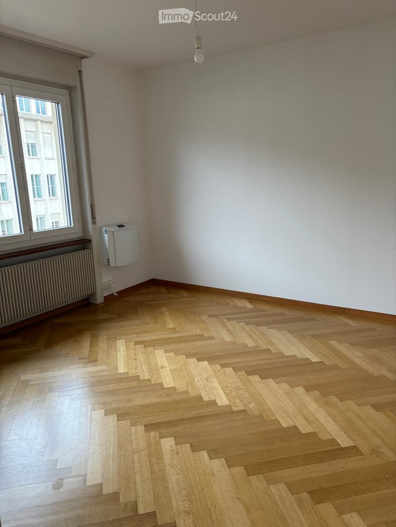 Empty room with wooden herringbone flooring, white walls, large window with view, radiator, and a wall-mounted air conditioning unit.