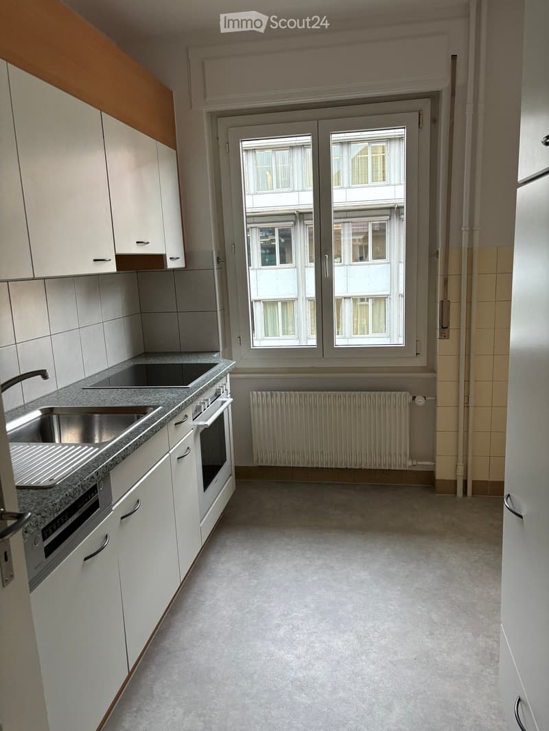 Modern kitchen with white cabinets, granite countertop, stainless steel sink, oven, and radiator. Large window with double doors offers a view of the building outside.