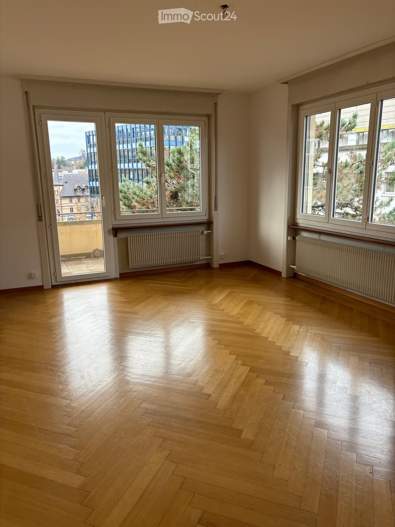 Empty room with wooden flooring, large windows, and radiators along the walls. The room has a sliding glass door leading to a balcony or outdoor area.