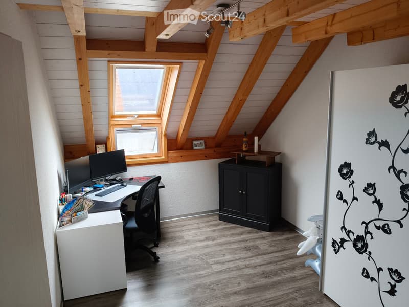 Cozy attic office with wooden beams, slanted ceilings, and large windows. Includes a desk with a computer, chair, cabinet, and decorative items.