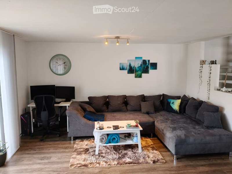 A modern living room with a grey sectional sofa, a white coffee table, a chair, a desk with a computer, and a clock on the wall.