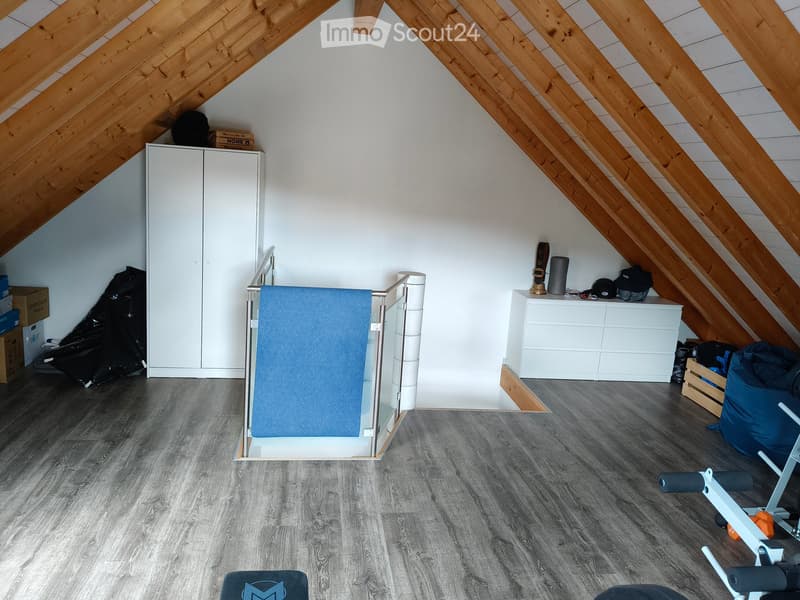 An attic room with wooden slanted ceilings, grey wooden flooring, a white cabinet, a mirror, and exercise equipment including a bike and a stool.